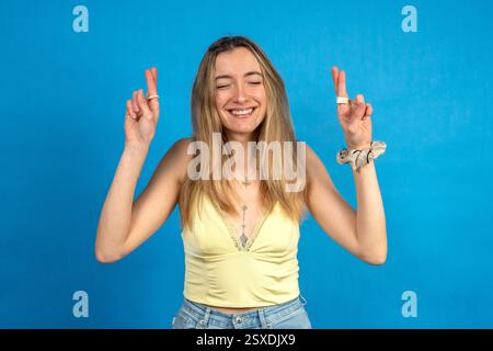 Portrait studio d'une jeune femme joyeuse avec les doigts croisés faisant un vœu avec les yeux fermés, espérant bonne chance Banque D'Images