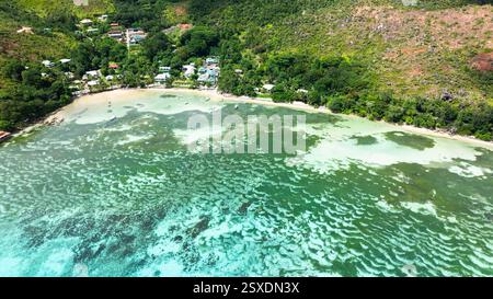 Vue aérienne des eaux peu profondes et claires près d'un village côtier, avec des collines verdoyantes s'élevant en arrière-plan. Praslin, Seychelles. Banque D'Images