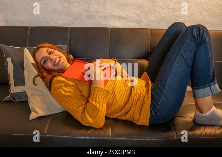 Heureuse jeune femme allongée sur un confortable canapé noir, serrant un livre rouge et souriant Banque D'Images
