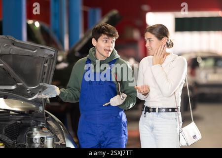 Mécanicien conseille jeune femme cliente sur la réparation sous le capot de la voiture dans la station-service de voiture Banque D'Images