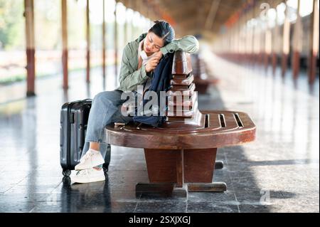 Une femme asiatique fait une sieste en étant assise les jambes croisées sur un banc en bois avec son sac et ses bagages dans la gare ou la gare. Banque D'Images