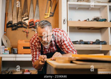Un menuisier senior en chemise à carreaux rouge sable soigneusement un morceau de bois dans son atelier. Les outils pendent Banque D'Images