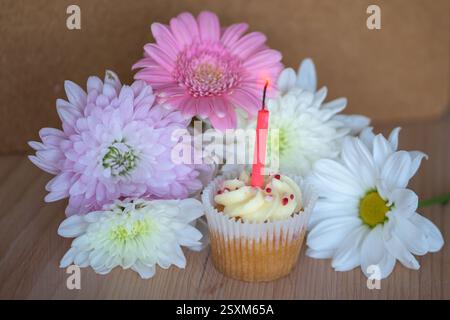 Cupcake coloré avec bougie entouré de fleurs vibrantes sur la surface en bois, ambiance chaleureuse, festive et festive. Banque D'Images