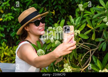 Une jeune femme souriante portant un chapeau de soleil et des lunettes de soleil, entourée de buissons de fleurs en fleurs, prend un selfie avec son téléphone portable. Banque D'Images