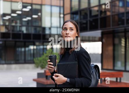 Jeune femme entrepreneure tenant le café pour aller porter des vêtements noirs. Élégante femme se tient debout dans la cour d'un immeuble de bureaux, tenant une digi Banque D'Images