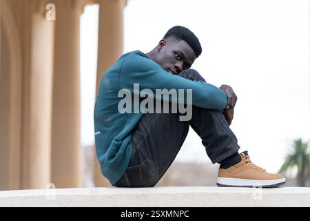 Jeune étudiant afro-américain se sentant seul et déprimé, serrant ses genoux tout en étant assis sur un mur Banque D'Images