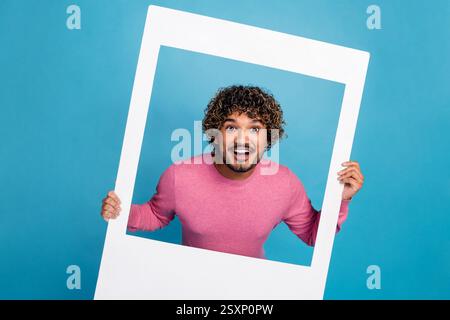 Jeune homme joyeux dans un pull rose décontracté tenant un cadre blanc sur un fond bleu, exprimant le bonheur Banque D'Images