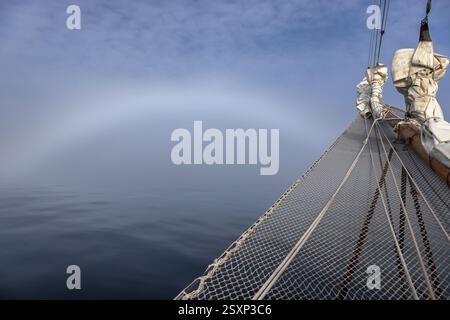 Proue d'un voilier coupant dans le brouillard à Hall Bredning, Scoresby Sund, Groenland. Il y a un arc de brouillard dans le ciel, un phénomène causé par la lueur Banque D'Images