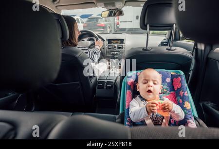 Portrait de bébé riant dans un siège d'auto tandis que mère conduisant une voiture de tourisme. Scène capture le moment de soin et de responsabilité, soulignant l'importance Banque D'Images