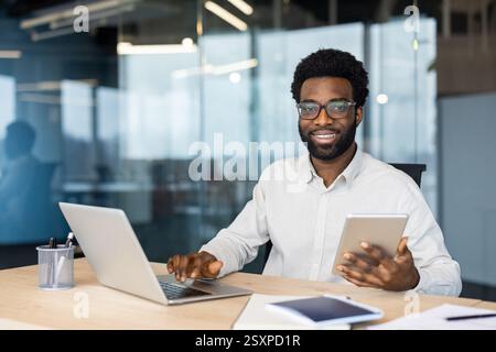Portrait d'un homme d'affaires prospère à l'intérieur du bureau sur son lieu de travail. L'homme sourit et regarde la caméra, en utilisant une tablette Banque D'Images
