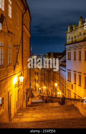 Le charmant escalier menant au château de Prague est baigné de lumière jaune chaude. Les bâtiments environnants créent une atmosphère chaleureuse, tandis que le ciel nocturne ajoute de la profondeur à la scène. Banque D'Images
