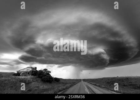 Paysage noir et blanc spectaculaire avec une tempête supercellulaire qui menace une ferme abandonnée et une route de campagne tranquille Banque D'Images