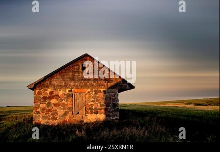 Vieilles grange en pierre et en bois qui résistent aux éléments dans un champ de blé baigné par la lueur chaude du coucher du soleil Banque D'Images