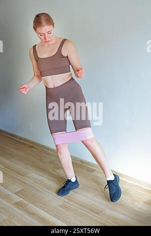 Femme faisant de l'exercice de squat avec bande de résistance à la maison, portant des vêtements de sport, en se concentrant sur la forme physique Banque D'Images