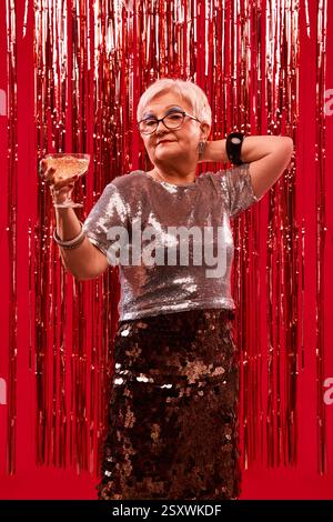 Femme âgée en haut argenté et jupe pétillante tenant un verre de champagne sur fond rouge scintillant. Style de vie glamour et ambiance de fête. Banque D'Images