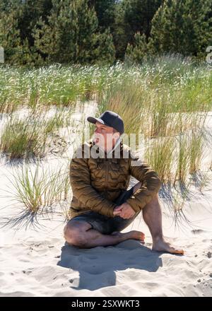 Homme d'âge moyen en veste et short est assis sur le sable de la mer Baltique. Des dunes de sable mou peuvent être vues en arrière-plan. Tranquillité, réflexion et contemplation sur Banque D'Images