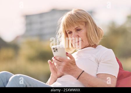 Assis sur le fauteuil et à l'aide d'un smartphone. Femme senior ayant un bon week-end à l'extérieur sur le terrain à la journée ensoleillée. Banque D'Images