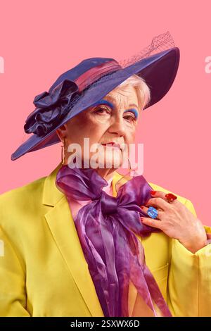 Femme âgée en blazer jaune portant un chapeau bleu regardant en toute confiance tout en ajustant le grand arc violet sur fond rose. Banque D'Images