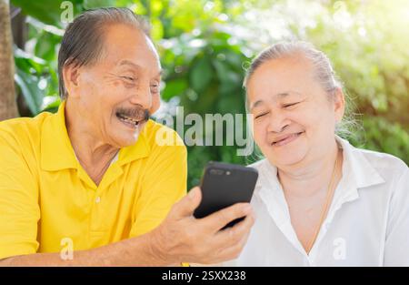 Heureux couple de personnes âgées asiatiques riant assis sur une chaise de jardin tout en utilisant la technologie intelligente dans le jardin. Concept extérieur. Banque D'Images