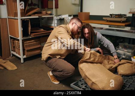 Plan à angle élevé de deux hommes caucasiens agenouillés près d'un énorme sac plein de nouveaux grains de café, ils l'ouvrent soigneusement pour vérifier le produit Banque D'Images