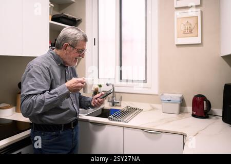 Homme senior appréciant le café et utilisant un smartphone dans la cuisine moderne Banque D'Images