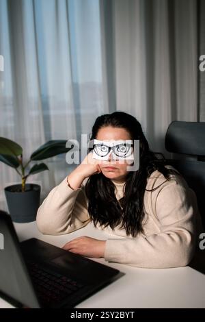 Employée de bureau épuisée et fatiguée femme avec les yeux peints sur des autocollants dormant sur le lieu de travail. Femme n'ayant pas d'énergie pour le travail assis au bureau avec adh Banque D'Images