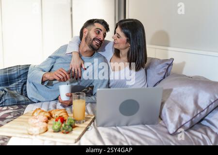 Jeune couple se relaxant au lit, prenant le petit déjeuner et regarder un film sur leur ordinateur portable, profitant d'une matinée confortable ensemble Banque D'Images