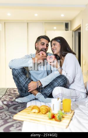 Jeune couple portant un pyjama, assis dans le lit et mangeant le petit déjeuner, y compris des beignets, des croissants, du jus d'orange et des fraises Banque D'Images