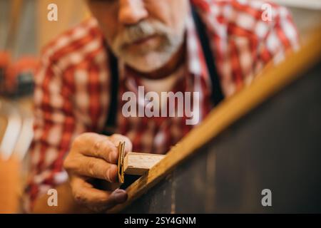 Un menuisier senior en chemise à carreaux rouge sable soigneusement un morceau de bois dans son atelier. Les outils pendent Banque D'Images