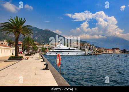 Un yacht de luxe est amarré à Tivat, au Monténégro, entouré de palmiers, de bâtiments élégants et de montagnes pittoresques sous un ciel lumineux et partiellement nuageux Banque D'Images