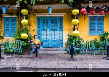 Une femme est assise sur une chaise devant une maison colorée décorée de lanternes dans la vieille ville de Hoi an, Hội an. Banque D'Images