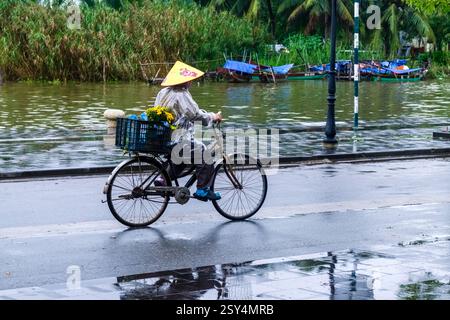 Une femme avec un chapeau conique sur une bicyclette roule sur une route sur la rive de la rivière Thu bon à Sông Thu Bồn un jour de pluie. Banque D'Images