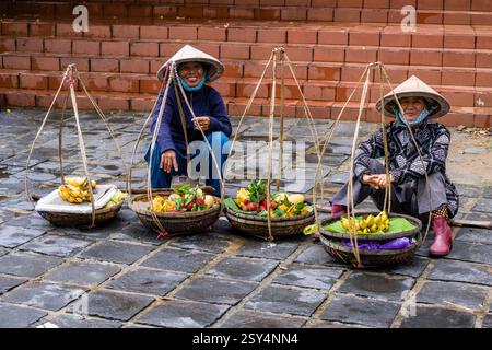 Deux femmes portant des chapeaux coniques vendent différents types de fruits dans des paniers dans les rues du centre de Hoi an, Hội an. Banque D'Images
