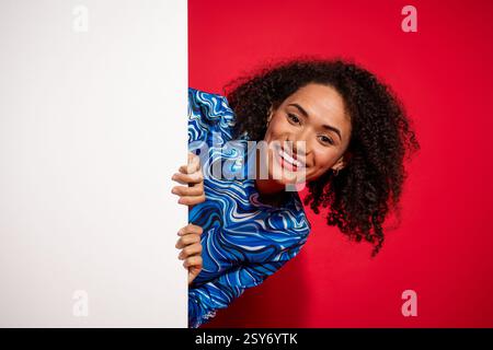 Jeune femme joyeuse avec des cheveux bouclés élégants se penchant pour jeter un coup d'œil derrière un panneau blanc, portant un haut imprimé coloré Banque D'Images