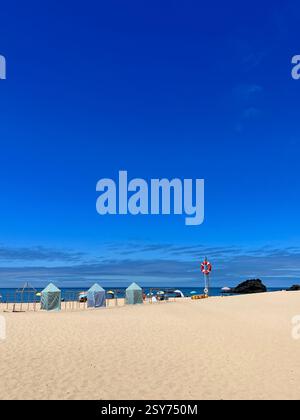 Une plage paisible avec des tentes rayées, des parasols et une bouée de sauvetage sous un ciel bleu clair. Banque D'Images