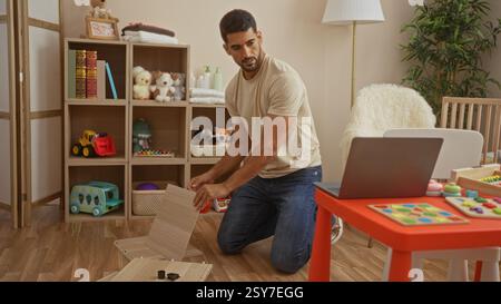 Bel homme assemblant des meubles dans une chambre confortable avec des jouets et un berceau, entouré de livres, de plantes et d'un ordinateur portable, indiquant un environnement familial chaleureux Banque D'Images
