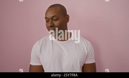 Jeune homme sur fond rose isolé regarde vers le bas avec un sourire subtil, portant un t-shirt blanc décontracté, créant une atmosphère vibrante et moderne avec Banque D'Images