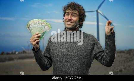 Homme hispanique tenant des roubles russes dans un champ avec des moulins à vent, faisant un geste suggestif de richesse et de prospérité. Banque D'Images