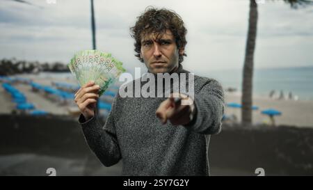 Homme hispanique sur la plage de bord de mer pointant vers la caméra tenant des roubles russes, combinant motivation financière et détente en plein air. Banque D'Images