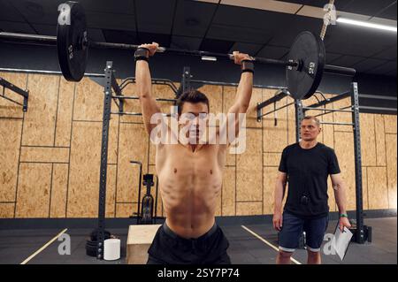 Jeune homme musclé soulevant la barre dans le gymnase. Formateur debout et regardant. Banque D'Images