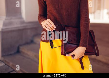 Femme à la mode portant un pull en cachemire marron et une jupe jaune tenue dans la rue de la ville avec sac à main en cuir élégant tenant des lunettes de soleil Banque D'Images