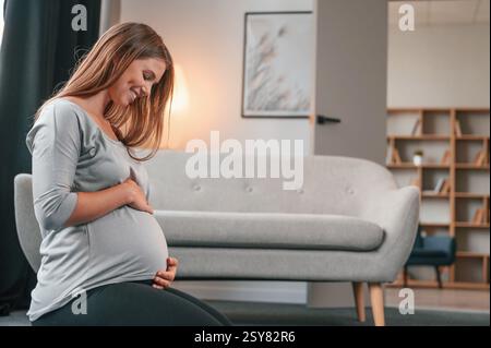 Vue latérale. Assis sur le sol. Belle femme enceinte est à l'intérieur à la maison. Banque D'Images