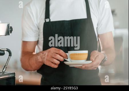 Vue rapprochée. Tenir une tasse de café. Travailleur de café en chemise blanche et tablier noir est à l'intérieur. Banque D'Images