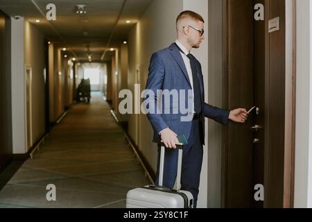 Gros plan moyen d'un homme qui vient d'arriver avec une valise argentée debout devant la porte en utilisant une carte pour ouvrir sa chambre d'hôtel Banque D'Images