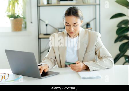 Concentrée femme d'affaires prospère confiante, tenant une carte de crédit et utilisant un ordinateur portable tout en étant assise à un bureau dans son bureau moderne, accomplissant des tâches en ligne, faisant des paiements en ligne, entrant la carte Banque D'Images