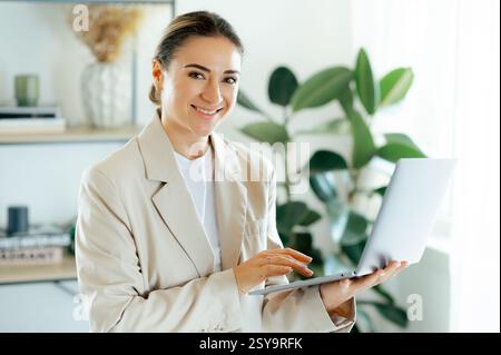 Professionnel charmante femme d'affaires caucasienne réussie dans un costume beige tenant un ouvert dans les mains, debout dans son espace de bureau tout en posant avec confiance pour la caméra, sourire amical, prêt pour le travail Banque D'Images