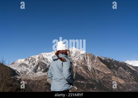 Vue arrière d'une jeune femme méconnaissable dans un chapeau d'hiver et des stands de veste face à une vue imprenable sur les montagnes enneigées, incarnant une rura sereine Banque D'Images