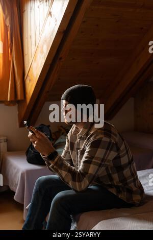Un homme est capturé à l'aide de son smartphone alors qu'il est assis confortablement sur un lit dans un grenier. Il est vêtu chaudement d'un bonnet et d'une chemise à carreaux, reflétant un Banque D'Images