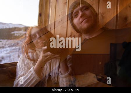Une scène hivernale confortable capture une femme rousse tenant un appareil photo, reflétée aux côtés d'un homme dans une fenêtre en verre, sur fond de neige Banque D'Images