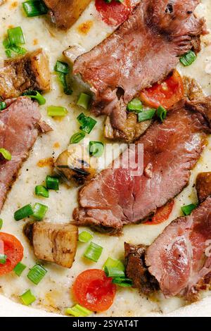 Vue rapprochée sur une pizza à la mozzarella garnie de rôti de bœuf, de champignons porcini, de tomates cerises et saupoudrée d'oignons verts. Banque D'Images